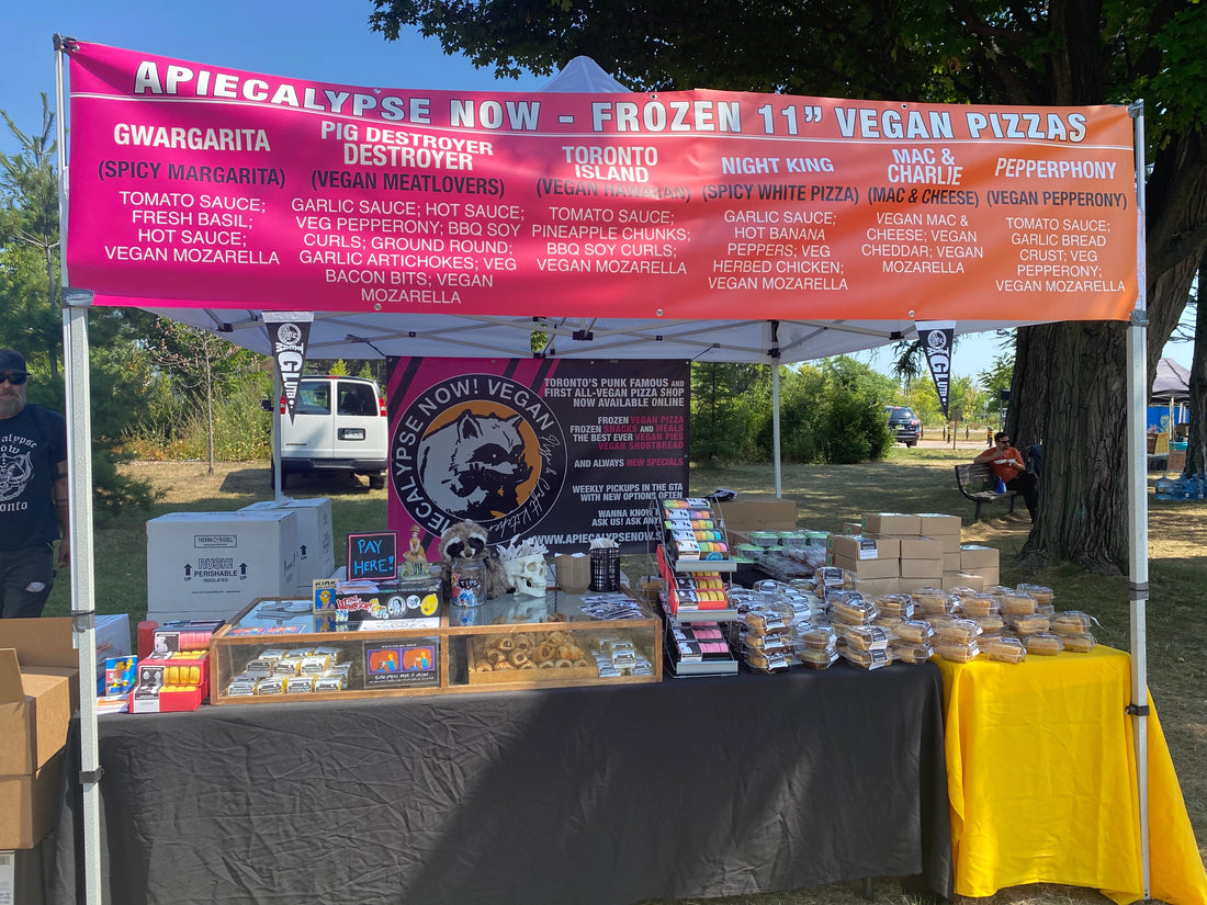 A photograph of our veg fest booth at Burlington Veg Fest this past summer. A banner advertising our pizzas, a table full of pies and treats, black tablecloth, and our raccoon banner. 